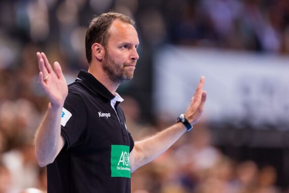 Ein Mann in einem schwarzen Polohemd hebt beide Hände, die Handflächen nach außen, als wolle er während einer Sportveranstaltung ein Zeichen geben oder kommunizieren. Der Hintergrund ist mit Zuschauern unscharf.