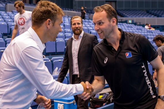 Zwei Männer, einer in einem weißen Hemd und einer in einem schwarzen HANDBALL-Polo, geben sich in einer Sportarena die Hand und lächeln, während andere im Hintergrund stehen und zusehen.