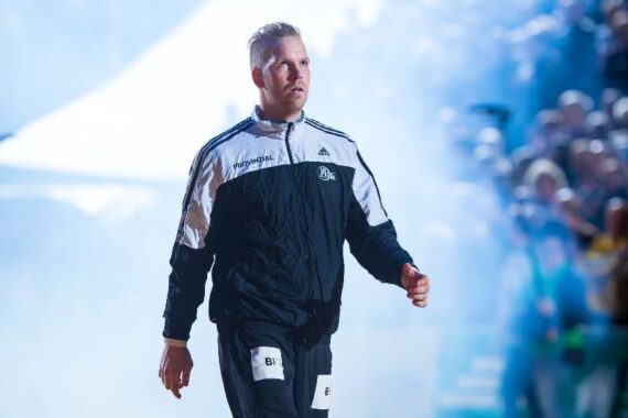 Ein männlicher Sportler in einem schwarz-weißen Trainingsanzug geht selbstbewusst an einer jubelnden Menge und hellen Lichtern vorbei, mit Rauch oder Nebel im Hintergrund.