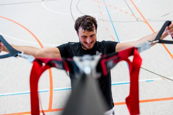 Ein Mann in einer Sporthalle lächelt, während er sich an den Griffen roter Gymnastikbänder festhält, während unter ihm auf dem Boden verschiedene farbige Spielfeldlinien zu sehen sind.