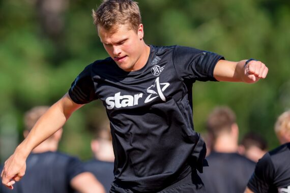 Ein junger Mann, der ein schwarzes Sporthemd mit aufgedrucktem Stern trägt, befindet sich während einer sportlichen Trainingseinheit im Freien mitten im Sprung, mit unscharfen Mannschaftskameraden und Grünflächen im Hintergrund.