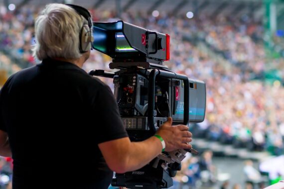 Ein Kameramann mit Kopfhörern bedient eine große Videokamera auf einem Stativ und filmt eine Veranstaltung in einer überfüllten Halle mit vielen unscharfen Zuschauern im Hintergrund.
