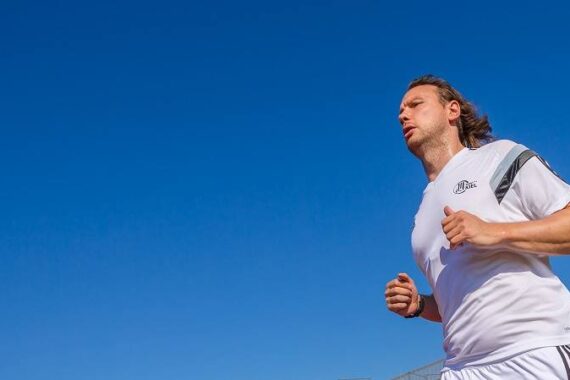 Ein Mann in weißer Sportkleidung joggt im Freien unter einem klaren blauen Himmel. Er wirkt konzentriert und ist aus einem niedrigen Winkel aufgenommen, der den offenen Himmel über ihm hervorhebt.