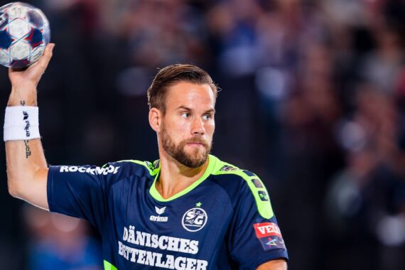 Ein männlicher Handballspieler mit Bart, der ein marineblaues und neongrünes Trikot trägt, hält einen Handball über seine Schulter und bereitet sich auf den Wurf vor. Der Hintergrund ist unscharf, um den Fokus auf den Sportler zu legen.