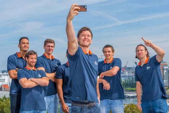 Sechs Männer in passenden blauen Polohemden posieren im Freien für ein Gruppen-Selfie, lächelnd und dicht beieinander stehend vor einer Stadtlandschaft mit Gebäuden und einem blauen Himmel im Hintergrund.