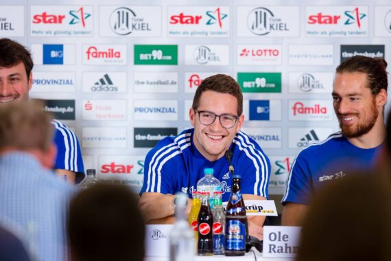 Drei lächelnde Männer in blauen Sporttrikots sitzen an einem Pressekonferenztisch mit Mikrofonen und Getränken, vor einem Hintergrund mit verschiedenen Sponsorenlogos. Der Mann in der Mitte spricht, und die Stimmung wirkt freundlich und entspannt.