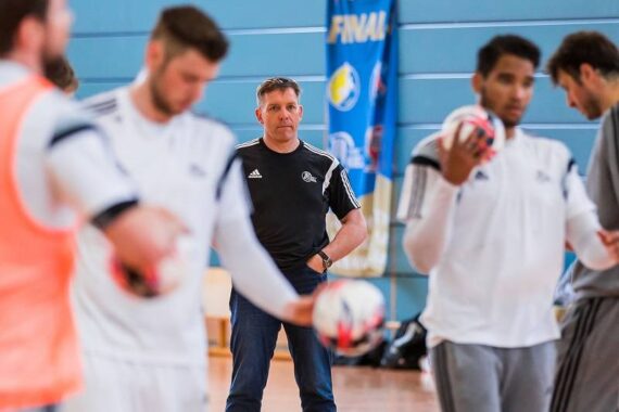 Ein Trainer in einem schwarzen Hemd sieht zu, wie mehrere Sportler in weißer Sportkleidung in einer Turnhalle mit Handbällen üben. Im Hintergrund sind blaue Wände und ein Transparent zu sehen.