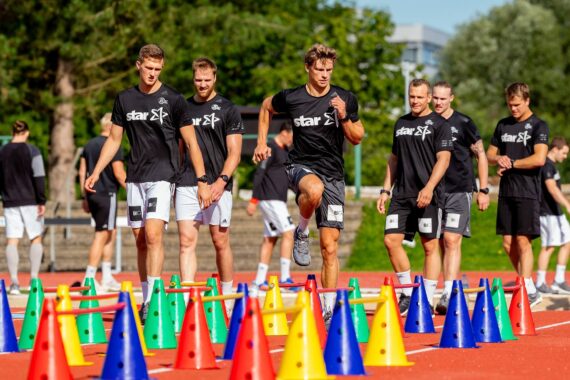 Eine Gruppe sportlicher Männer in passenden schwarzen Hemden und grauen Shorts trainiert im Freien, wobei ein Mann mitten im Sprung über bunte Kegel auf einer Bahn steht, während andere zusehen oder warten, bis sie an der Reihe sind. Im Hintergrund sind Bäume und Grünflächen zu sehen.