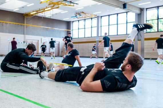 Mehrere Männer in Sportkleidung dehnen sich auf dem Boden einer Turnhalle, während andere im Hintergrund Handballübungen machen. Helles natürliches Licht erfüllt den Raum durch große Fenster.
