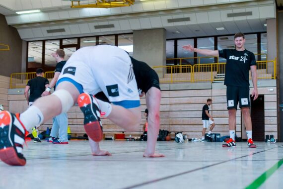 Zwei Sportler üben in einer Turnhalle Übungen. Einer ist in Bewegung und balanciert auf Händen und Füßen, während der andere steht, auf etwas zeigt und Sportkleidung trägt. Im Hintergrund sind Sportgeräte und Bänke zu sehen.
