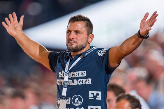 Ein Mann mit gestutztem Bart und kurzen Haaren trägt ein marineblaues Hemd mit weißer Aufschrift und steht mit erhobenen Armen, als wolle er feiern oder ermutigen. Im Hintergrund sind eine unscharfe Menschenmenge und der Veranstaltungsort zu sehen.