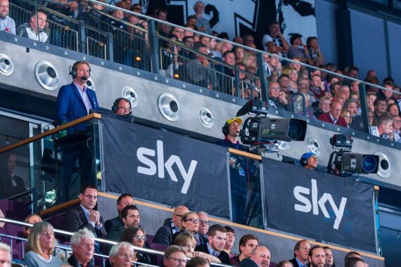 Fernsehkommentatoren in einer Kabine mit großen "Sky"-Bannern, umgeben von Kameras, überblicken eine überfüllte Arena mit Zuschauern bei einem Sportereignis.