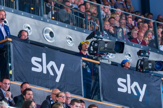 Fernsehteams und Kameraleute arbeiten in einer Stadionpressebox mit großen "Sky"-Bannern, während im Hintergrund eine Zuschauermenge sitzt und ein Ereignis verfolgt.