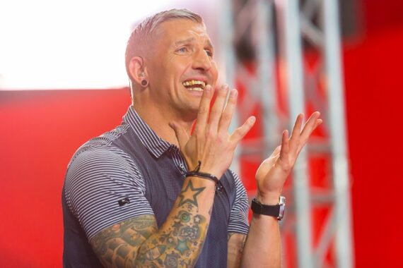Ein Mann mit tätowierten Armen und kurzen Haaren lächelt breit und hebt aufgeregt oder überrascht die Hände. Er trägt ein gestreiftes Poloshirt und steht in einem Innenraum vor einem verschwommenen rot-weißen Hintergrund.