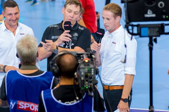 Drei Männer stehen zusammen und halten Mikrofone mit der Aufschrift Sky Sport. Sie sprechen und gestikulieren, während sie einem Kameramann während einer Sportveranstaltung in einer Halle gegenüberstehen.