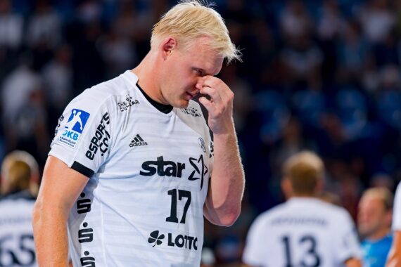 Ein blonder männlicher Handballspieler in einem weißen Trikot mit der Nummer 17 steht auf dem Spielfeld und berührt sein Gesicht mit einem müden oder enttäuschten Ausdruck. Im Hintergrund sind andere Spieler und eine verschwommene Menschenmenge zu sehen.
