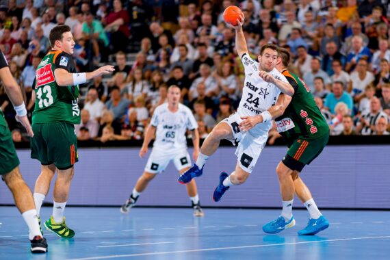 Ein Handballspieler in einem weißen Trikot springt, um den Ball zu werfen, der von Spielern in grünen Trikots während eines Spiels eng verteidigt wird. Das Spielfeld befindet sich in einer Halle mit einer jubelnden Menge im Hintergrund.
