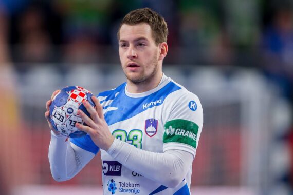 Ein männlicher Handballspieler in einem weiß-blauen Trikot hält einen Handball und bereitet sich auf den Wurf vor. Sein Trikot trägt die Nummer 23 und die slowenische Flagge. Der Hintergrund ist mit Zuschauern unscharf.
