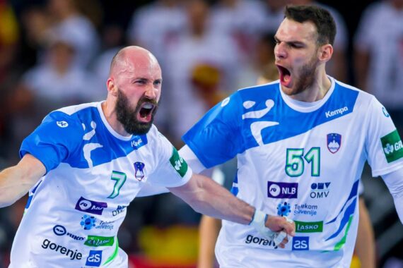 Zwei männliche Handballspieler in weißen und blauen Trikots feiern leidenschaftlich, mit aufgerissenem Mund und geballten Fäusten, die Aufregung und Intensität eines Spiels zeigen.
