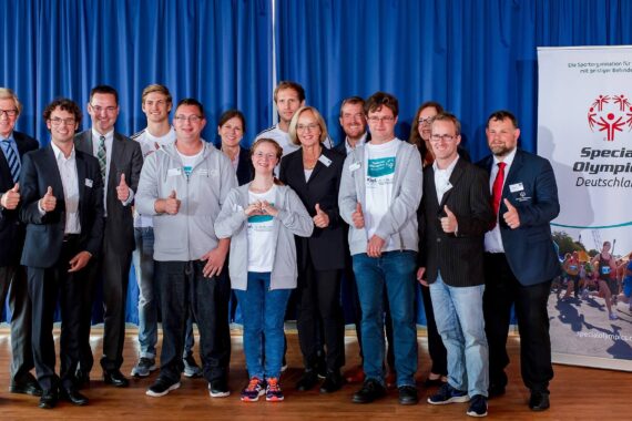 Eine Gruppe von Menschen, einige in Anzügen, andere in Freizeitkleidung, steht in einem Innenraum zusammen, lächelt und zeigt die Daumen nach oben, vor einem blauen Vorhang und einem Banner von Special Olympics Deutschland.