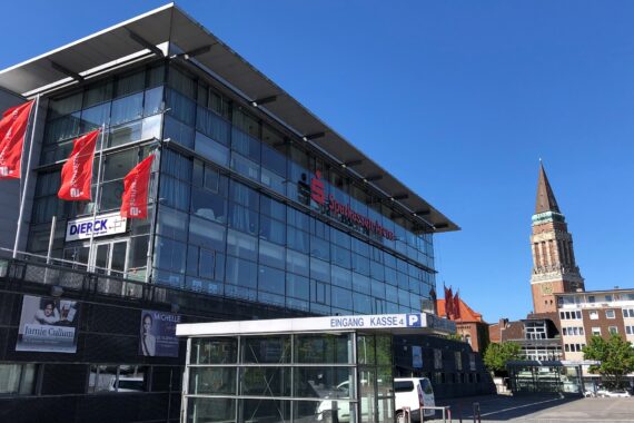 Ein modernes Glasgebäude mit roten Fahnen und dem Logo der Sparkassen-Arena, neben einer Parkeinfahrt. Im Hintergrund erhebt sich ein historischer Uhrenturm aus Backstein vor einem strahlend blauen Himmel.