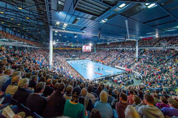 Eine große Halle voller Zuschauer, die ein Handballspiel auf einem hell erleuchteten Platz verfolgen, mit Spielern in Aktion und einer Anzeigetafel über der Mitte.