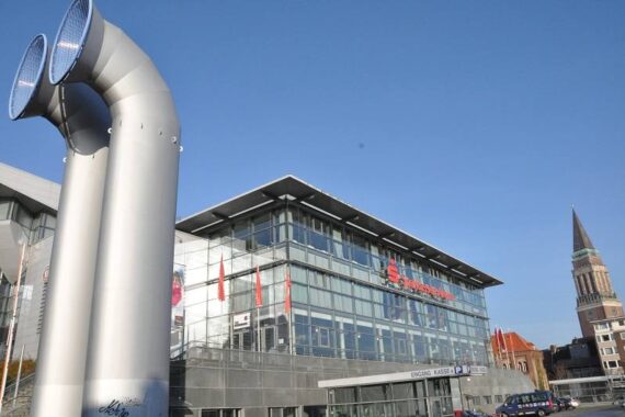 Modernes Glasgebäude mit dem Zeichen der Sparkassen-Arena, zwei große silberne Lüftungsrohre im Vordergrund, klarer blauer Himmel und ein hoher Kirchturm im Hintergrund.