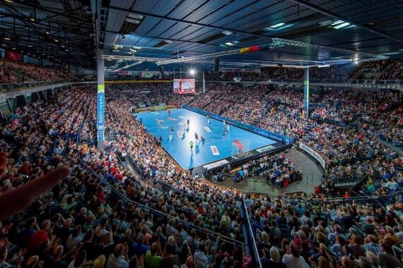 Eine überfüllte Halle mit Tausenden von Zuschauern, die ein professionelles Handballspiel auf einem hell erleuchteten blauen Spielfeld verfolgen, umgeben von gestaffelten Sitzplätzen unter einem großen Bogendach.