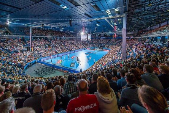 Eine große Halle voller Zuschauer, die ein Handballspiel auf einem hell erleuchteten blauen Spielfeld verfolgen. Die Tribünen sind voll, und die Zuschauer konzentrieren sich auf das Geschehen in der Mitte der Halle.