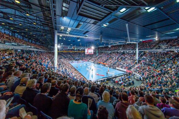 Eine große Halle, die mit Zuschauern gefüllt ist, die ein Handballspiel verfolgen. Das Spielfeld ist hell erleuchtet, während die Zuschauer auf stadionähnlichen Sitzen rund um das Spielfeld sitzen.