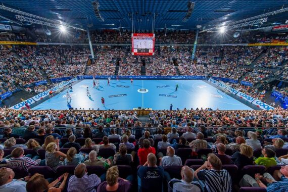 Eine große Halle, gefüllt mit Zuschauern, die ein Handballspiel auf einem blauen Spielfeld verfolgen, mit hellen Lichtern über dem Kopf und einer Anzeigetafel, die über der Mitte hängt.