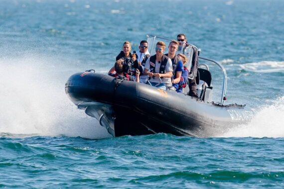 Eine Gruppe von sechs Personen in Schwimmwesten fährt in einem schnellen, schwarzen Schlauchboot über das kabbelige blaue Wasser und erzeugt weiße Gischt, während sie sich unter dem klaren Himmel vorwärts bewegen.