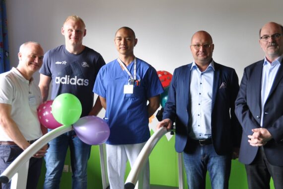 Fünf Männer stehen drinnen, einige lächelnd, vor einem Display mit bunten Luftballons. Einer trägt einen Arztkittel, zwei einen Anzug und zwei sind leger gekleidet. Im Hintergrund sind helle Vorhänge und Licht von einem Fenster zu sehen.