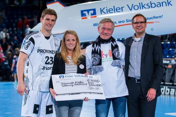 Vier Personen stehen auf einem Sportplatz und halten einen großen Spendenscheck der Kieler Volksbank in Höhe von 4.000 Euro in der Hand. Links ist ein Spieler im Sportdress zu sehen, im Hintergrund eine Flugzeugschraube.