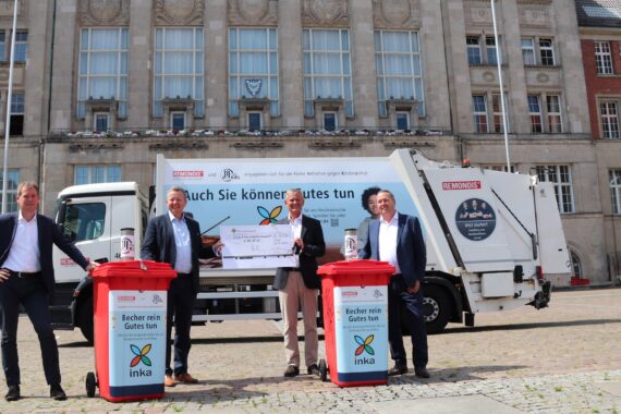 Vier Männer in Anzügen stehen vor einem Müllwagen, halten einen großen Scheck in der Hand und stehen neben zwei roten Recycling-Tonnen mit der Aufschrift inka. Die Szene spielt sich vor einem großen historischen Gebäude ab.