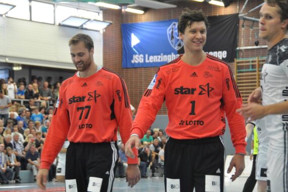 Zwei männliche Handballspieler in passenden orangefarbenen Trikots mit schwarzen Hosen stehen lächelnd auf einem Hallenplatz, im Hintergrund sitzt eine Menschenmenge und an der Wand hängt ein blaues Banner.