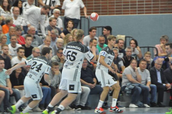 Drei Handballspieler in weißen Trikots laufen während eines Spiels auf einen Ball zu, während ein Schiedsrichter in Blau in der Nähe steht. Im Hintergrund ist eine Zuschauermenge zu sehen, die von der Tribüne aus aufmerksam zusieht.