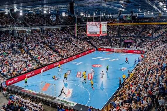 Eine große Halle, gefüllt mit Zuschauern, die ein professionelles Handballspiel auf einem blauen Spielfeld verfolgen, mit Spielern in Aktion und hellen Lichtern, die die Szene beleuchten.