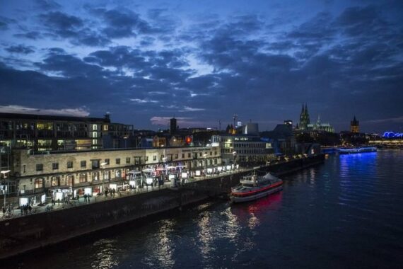 Eine Stadtlandschaft am Flussufer in der Abenddämmerung zeigt Gebäude mit beleuchteten Fenstern, ein angedocktes Boot auf dem Wasser und eine entfernte Kathedrale mit mehreren Türmen unter einem bewölkten Abendhimmel.