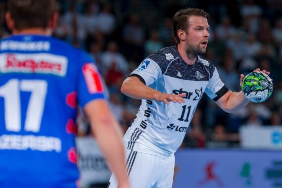 Ein Handballspieler in einem weiß-schwarzen Trikot hält einen grün-schwarzen Handball und bereitet sich auf den Wurf vor, während ein anderer Spieler in einem blauen Trikot mit der Nummer 17 von hinten zu sehen ist. Im Hintergrund ist eine unscharfe Menschenmenge zu sehen.