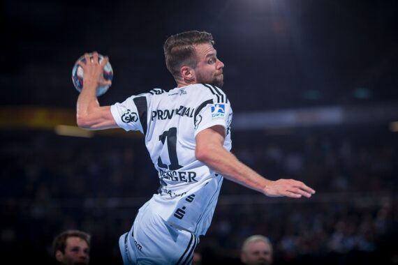 Ein männlicher Handballspieler in einem weiß-schwarzen Trikot springt in die Luft, um den Ball zu werfen. Der Hintergrund ist unscharf, um die Action und die Intensität des Spiels hervorzuheben.