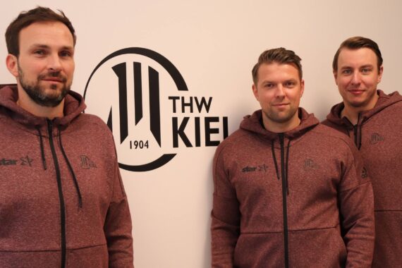 Drei Männer in passenden kastanienbraunen Kapuzenpullovern stehen vor einer weißen Wand mit dem Logo des Handballvereins THW Kiel und dem Aufdruck 1904. Sie schauen in die Kamera und stehen dicht beieinander.