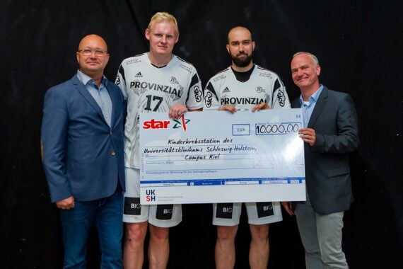 Vier Männer stehen zusammen und halten einen großen Spendenscheck in Höhe von 10.000 €, der an eine Kinderkrebsstation adressiert ist. Zwei Männer tragen Handballtrikots, die anderen Anzüge, alle lächeln vor einem schwarzen Hintergrund.