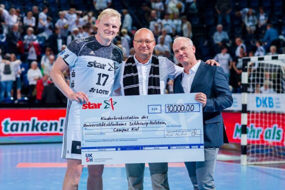 Drei Männer stehen in einer Sporthalle, lächeln und halten einen großen Scheck über 10.000 Euro in der Hand, der auf ein Kinderkrankenhaus ausgestellt ist. Ein Mann trägt ein Handballtrikot, die beiden anderen sind in Geschäftskleidung gekleidet.