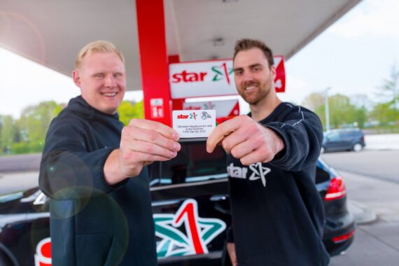 Zwei lächelnde Männer stehen an einer Tankstelle vor einem schwarzen Auto und halten eine Karte mit dem Stern-Logo in die Kamera. Beide tragen schwarze Hemden, und hinter ihnen ist der Schriftzug der Tankstelle zu sehen.