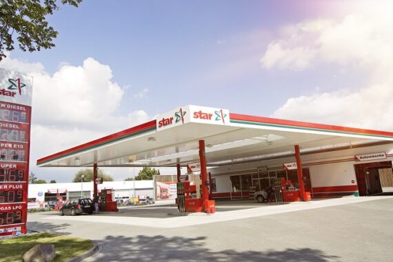 Eine moderne Star-Tankstelle mit einem großen Vordach, roten und weißen Akzenten, Zapfsäulen und einem Preisschild, das verschiedene Kraftstoffpreise anzeigt, unter einem teilweise bewölkten Himmel mit einfallendem Sonnenlicht.