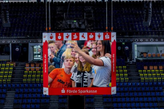 Eine Gruppe junger Leute macht ein Selfie, das auf einem großen Bildschirm in einer leeren Arena mit blauen und gelben Sitzreihen und roter Werbung um den Bildschirm herum angezeigt wird.