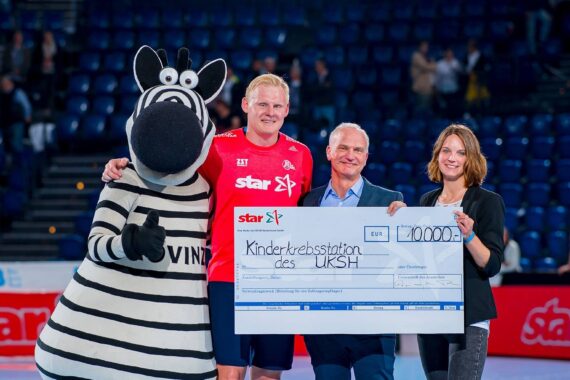 Vier Personen, darunter eine Person in einem Zebra-Maskottchen-Kostüm, stehen auf einem Sportplatz und halten einen großen Spendenscheck über 10.000 Euro in der Hand, während im Hintergrund Stadionplätze zu sehen sind.
