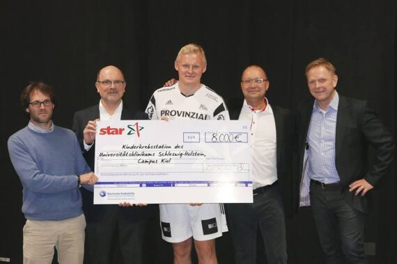 Fünf Männer stehen zusammen und halten einen großen Spendenscheck über 8.000 Euro, adressiert an das Universitätsklinikum Schleswig-Holstein, Camp Kiel. Ein Mann trägt eine Sportuniform, die anderen sind in Business-Kleidung gekleidet.