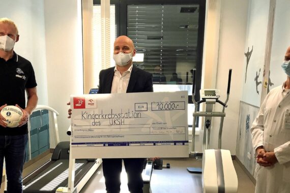 Drei Männer, alle mit Gesichtsmasken, stehen in einem Krankenzimmer. Einer hält einen Fußball in der Hand, ein anderer überreicht einen großen Spendenscheck über 10.000 Euro für eine Kinderkrebsstation, der dritte Mann trägt einen Arztkittel.